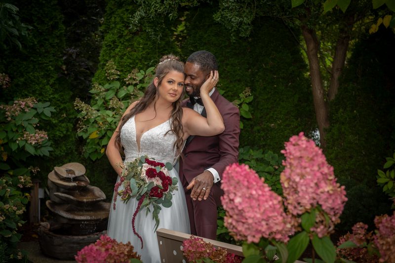 A bride in a white gown and floral bouquet stands close to her groom in a garden, hand resting on his cheek.