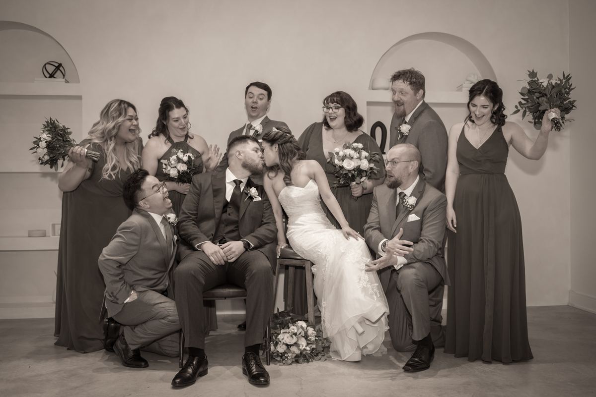 A bride and groom kiss joyfully as their wedding party reacts with laughter in a minimalist indoor setting.