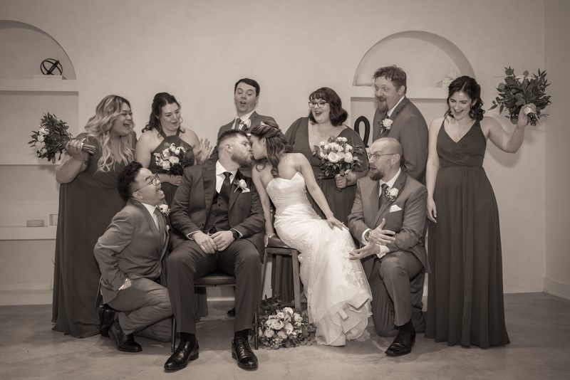 A bride and groom kiss joyfully as their wedding party reacts with laughter in a minimalist indoor setting.
