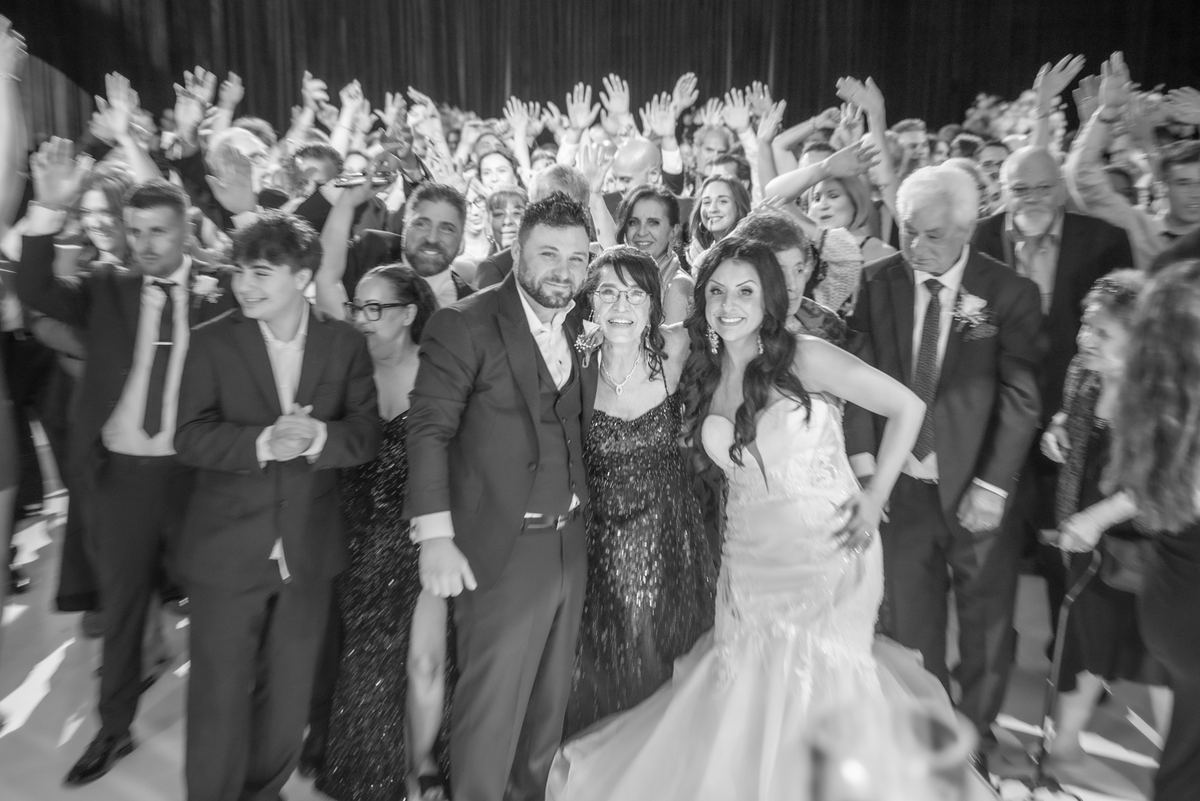 Bride and groom smile at center of dance floor surrounded by guests raising hands in celebration, conveying wedding joy.