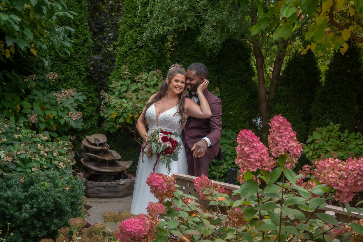 A bride and groom exchange a tender kiss in a garden filled with pink hydrangeas under diffused sunlight.