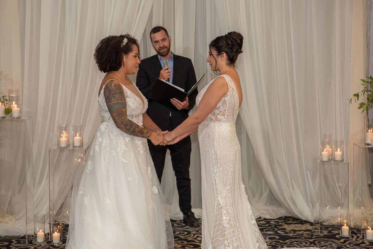Two brides exchange vows with an officiant between them, surrounded by lit candles and flowing white drapery.