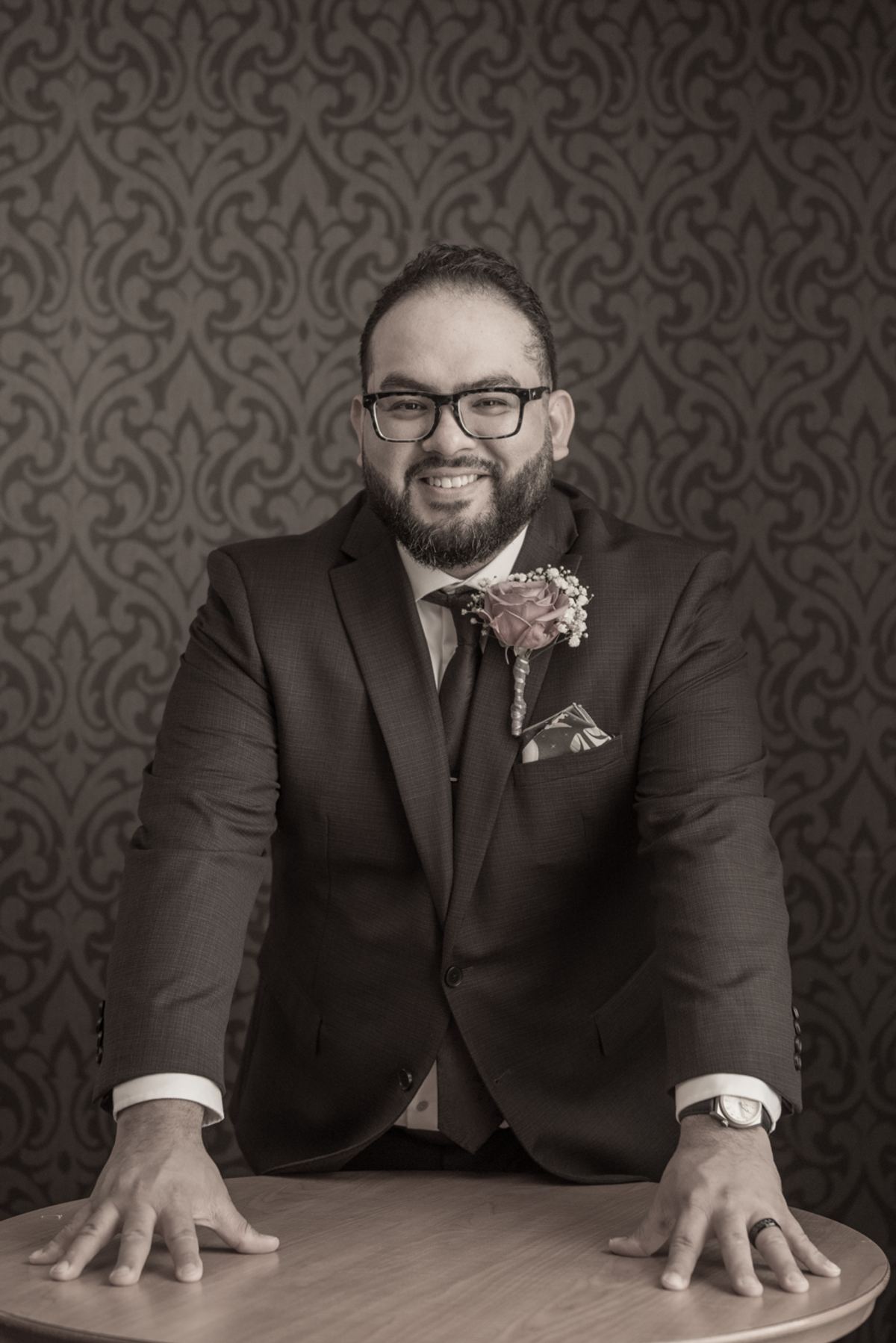 A groom in a suit poses confidently with his hands on a table, set against an ornate wallpaper backdrop.