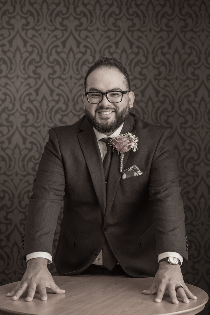 A groom in a suit poses confidently with his hands on a table, set against an ornate wallpaper backdrop.