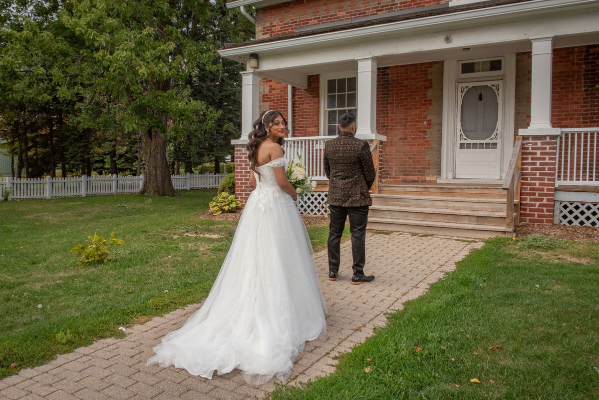 A bride in a white gown stands on a brick path, looking at her groom near a house with a porch, bathed in natural light.