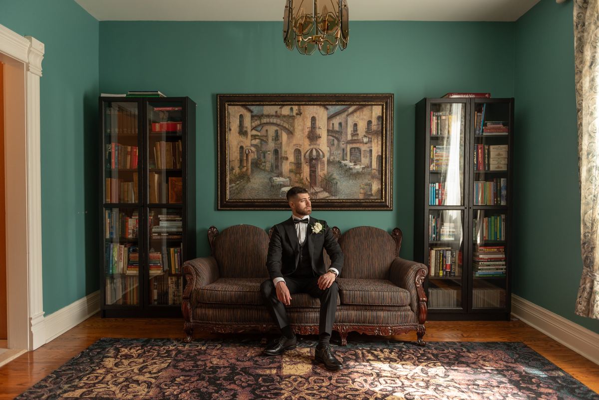 Groom in black tuxedo sits pensively on striped sofa in teal room with sunlight streaming through window.