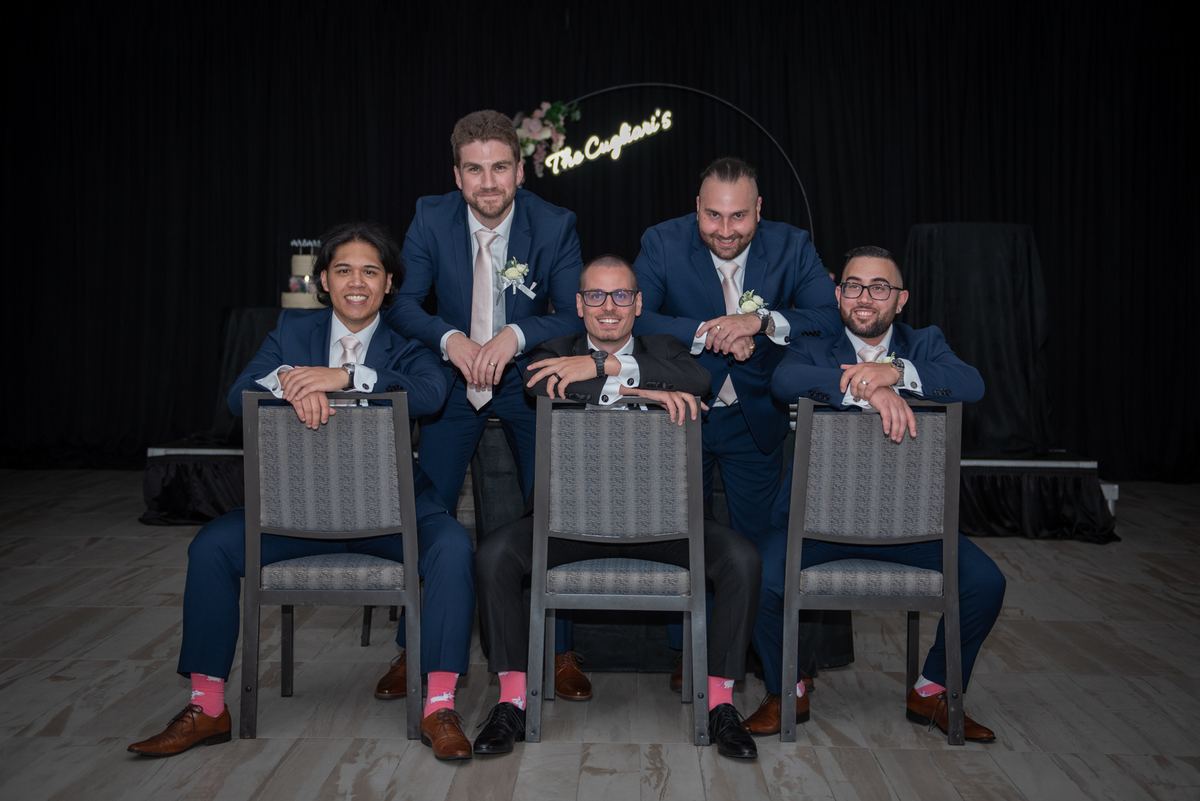 Five groomsmen and the groom pose in formal suits with matching boutonnieres against a dark backdrop, smiling.
