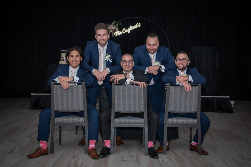 Five groomsmen and the groom pose in formal suits with matching boutonnieres against a dark backdrop, smiling.