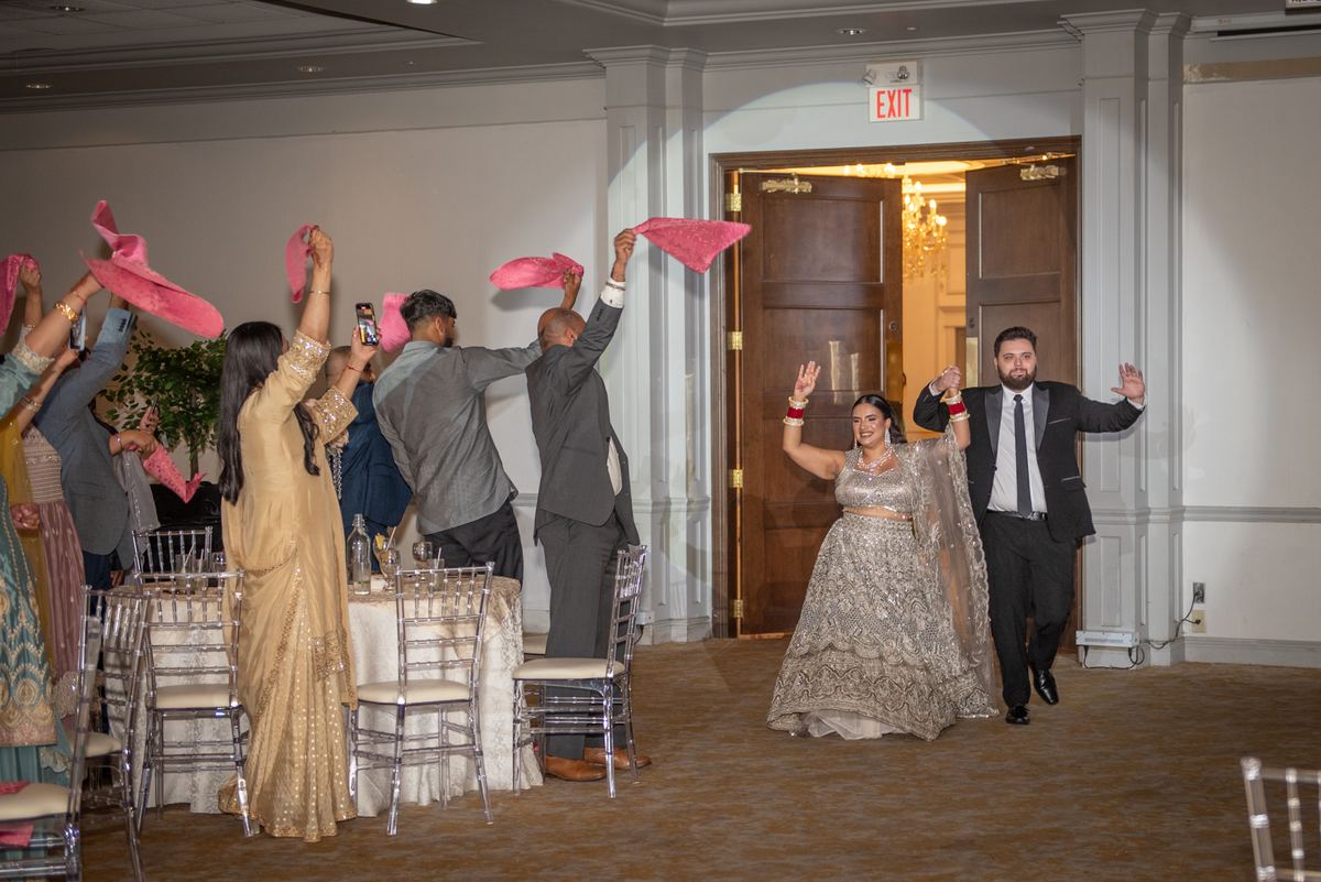 A bride and groom exit through a doorway into a celebration, greeted by waving guests with pink fabric scarves.