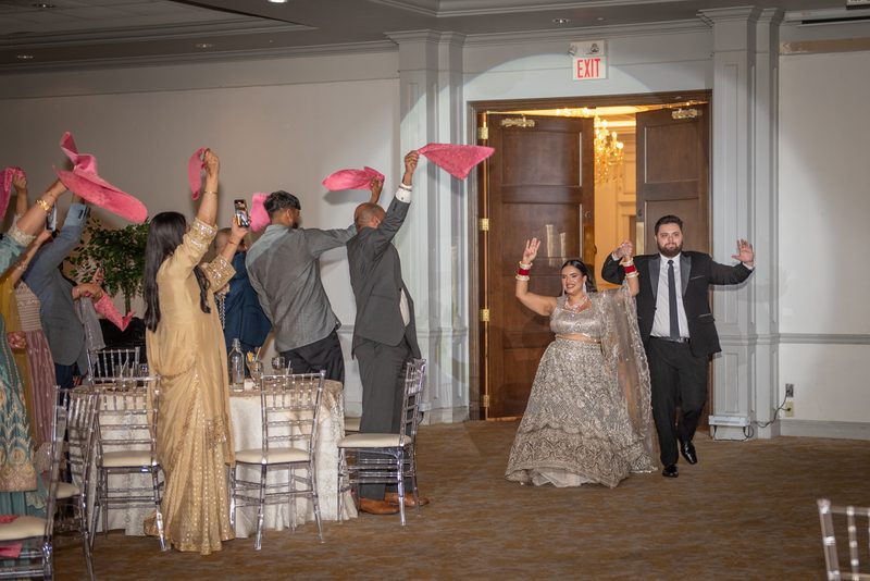 A bride and groom exit through a doorway into a celebration, greeted by waving guests with pink fabric scarves.