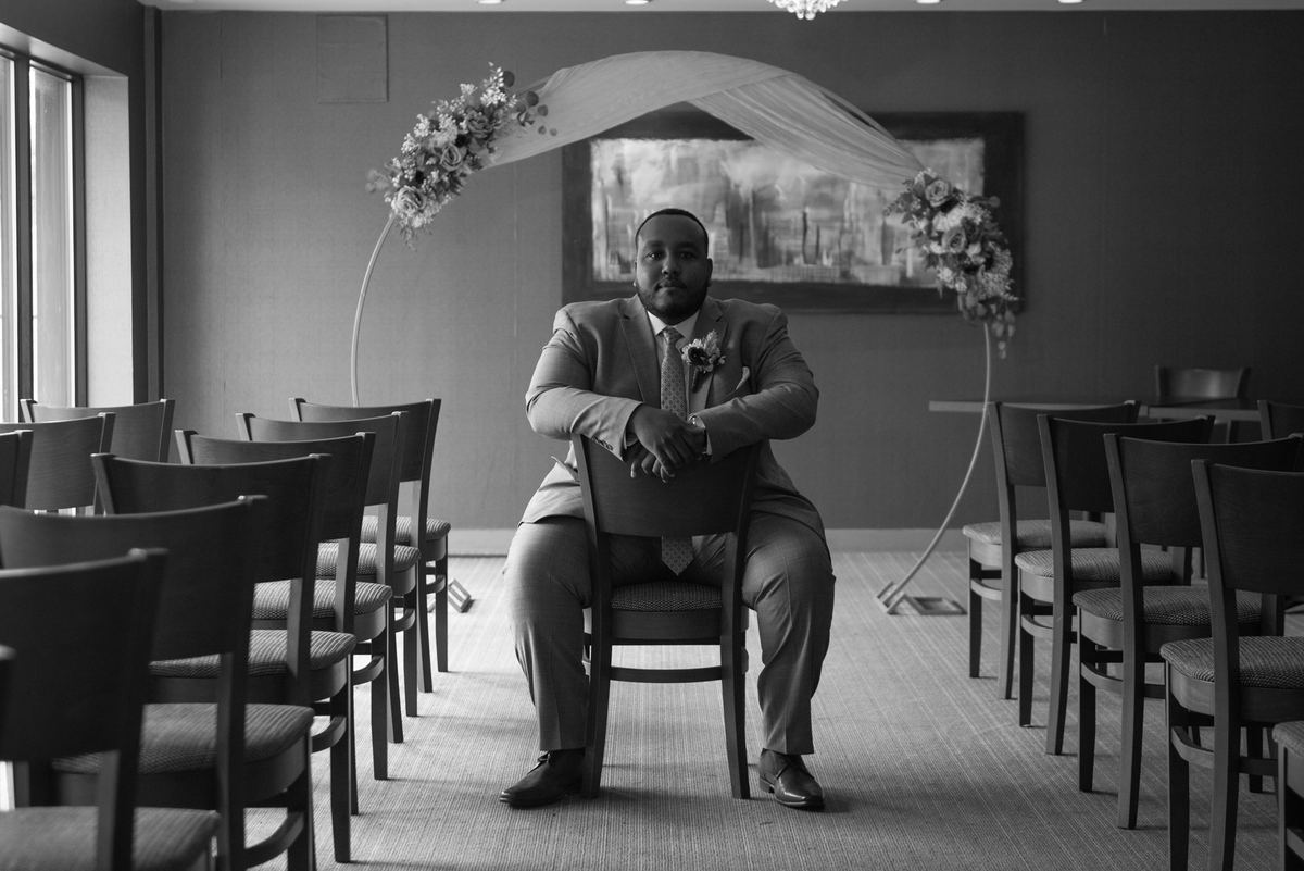 A groom in a suit and boutonnière sits quietly under a floral arch, gazing at the camera with composed intensity.