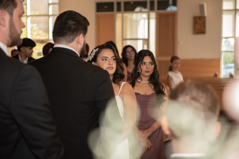 A bride in a white gown looks at her groom during their wedding ceremony inside a modern church, lit by natural light.