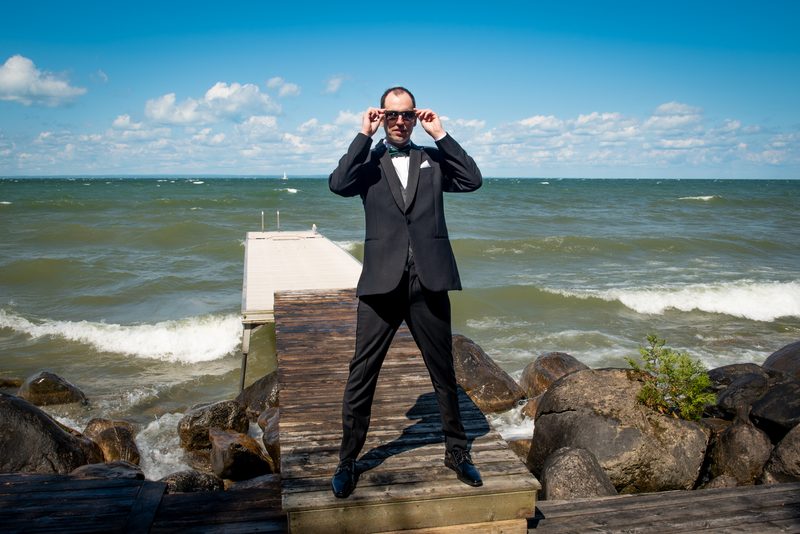 A man in a tuxedo stands on a pier by a rocky lakeshore, adjusting his sunglasses under bright skies.