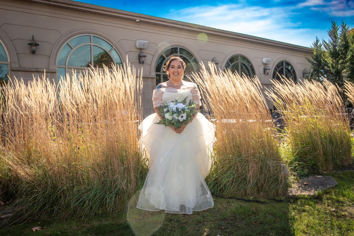A smiling bride in a white gown stands in a sunlit garden with tall golden grasses and an elegant building behind her.
