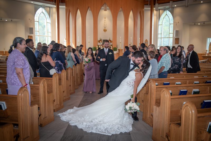 A bride and groom kiss at an altar, surrounded by wooden pews and smiling bridesmaids in mauve gowns.