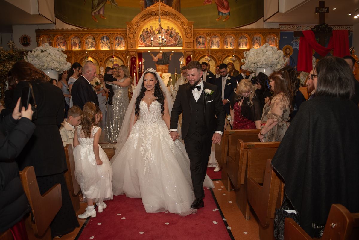 Bride and groom exit church aisle surrounded by guests and flowers, smiling as they walk past ornate gold iconostasis.