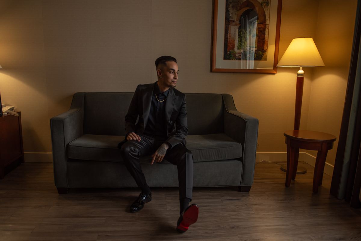 A man in a black suit sits thoughtfully on a gray sofa, his red shoe sole visible against the neutral tones of the room.