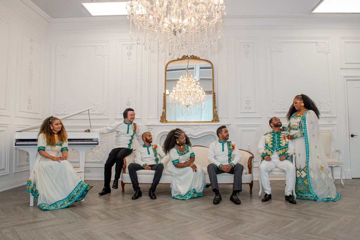 Seven elegantly dressed people pose around a piano and sofas in an ornate room under a chandelier, smiling.
