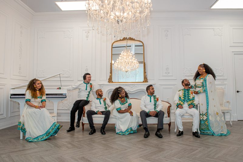 Seven elegantly dressed people pose around a piano and sofas in an ornate room under a chandelier, smiling.