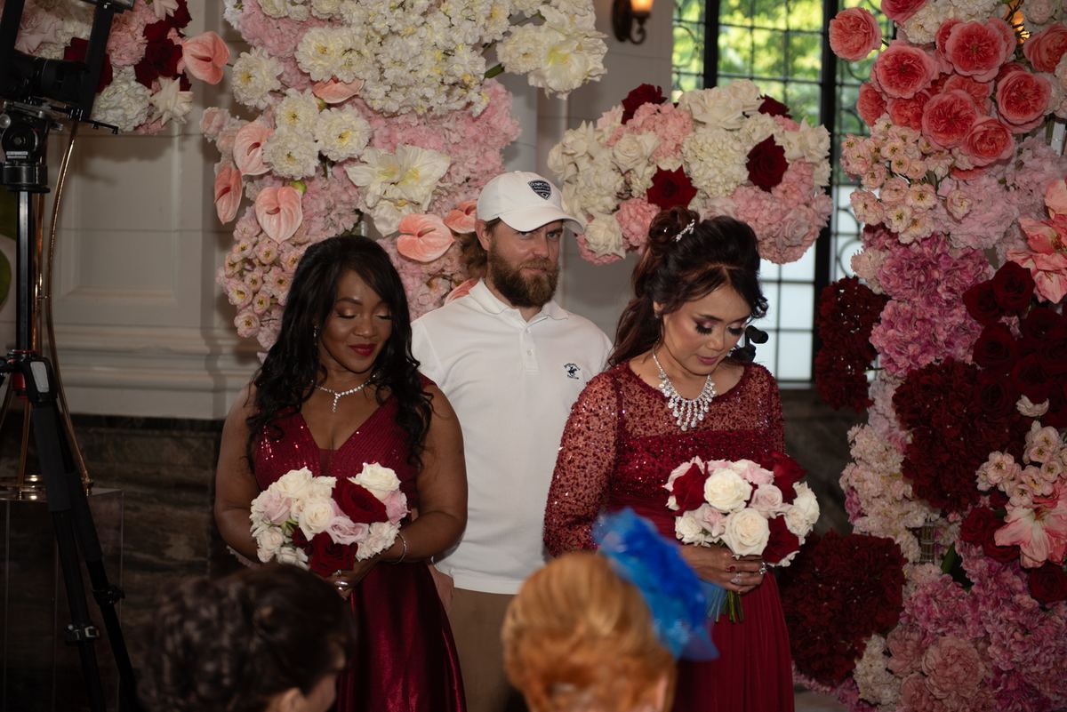 Two women in red dresses and a man in white stand before a floral backdrop, heads bowed in reverence during a ceremony.