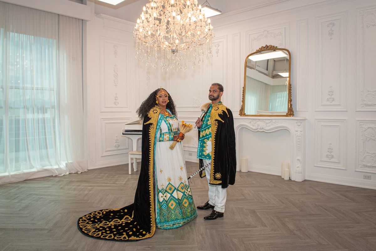 A couple in traditional gold and teal embroidered attire stand formally in a room with a chandelier.