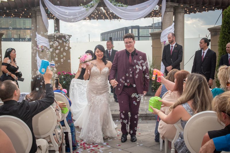 A bride and groom walk down the aisle under a decorated canopy, surrounded by guests blowing bubbles.