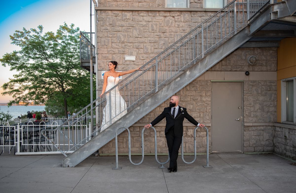 A bride and groom pose dramatically on a metal staircase near bike racks, with the waterfront visible in the background.