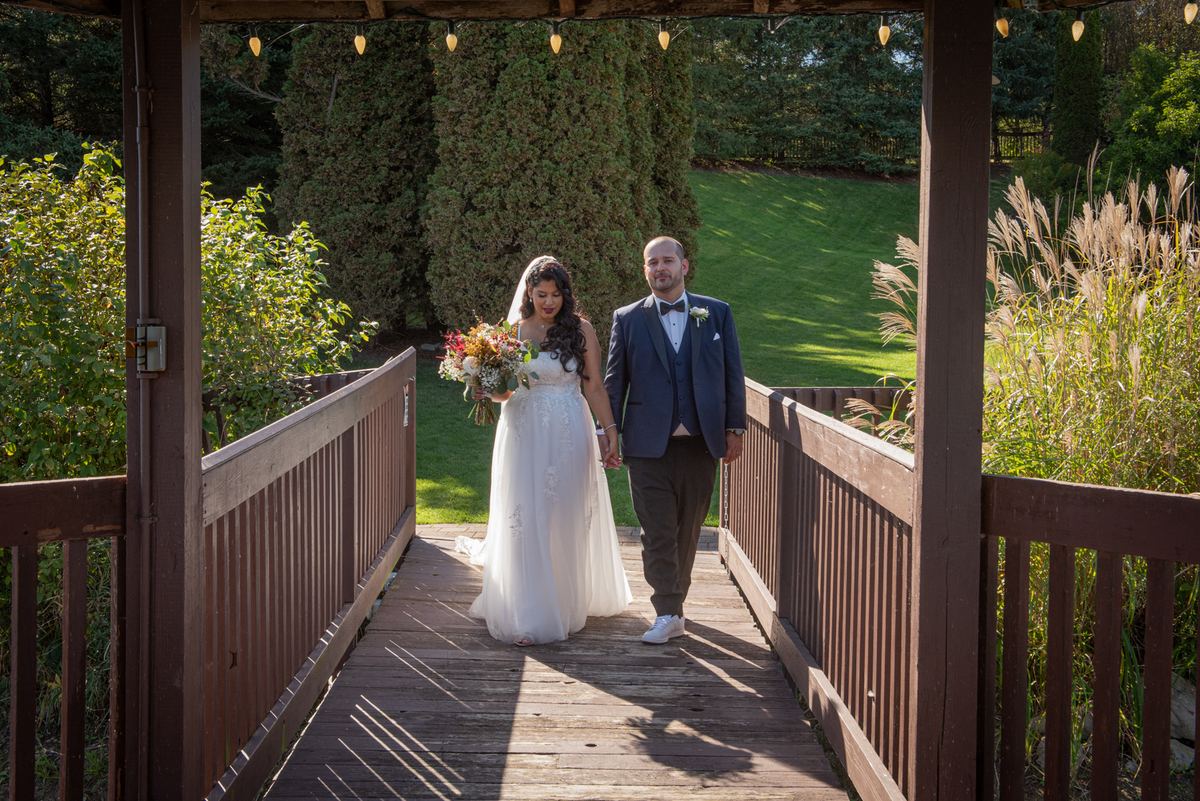 A bride and groom walk hand-in-hand across a wooden bridge, surrounded by string lights and framed by the structure's posts.