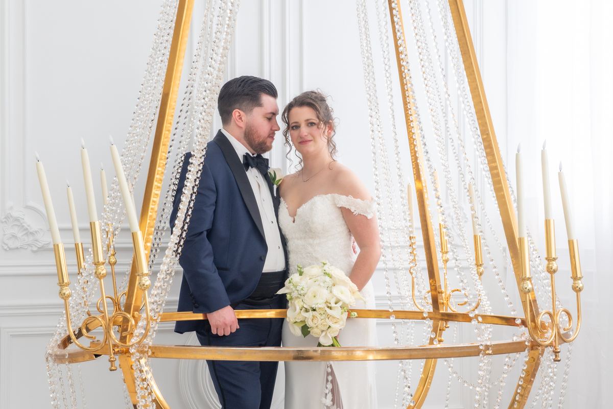 A newlywed couple stands closely beneath an ornate golden chandelier, illuminated by soft light and candles.