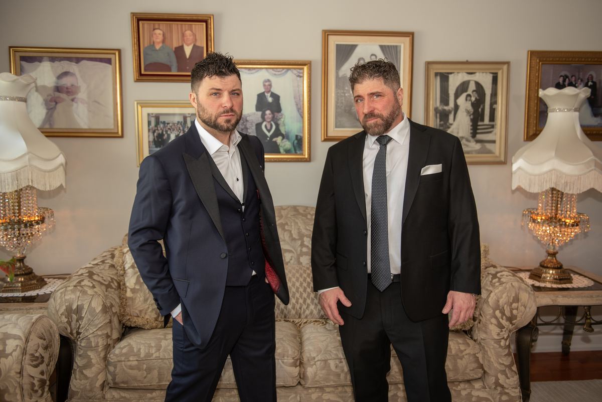 Two men in formal suits stand in an elegantly furnished room with family photos and ornate lamps, bathed in soft light.
