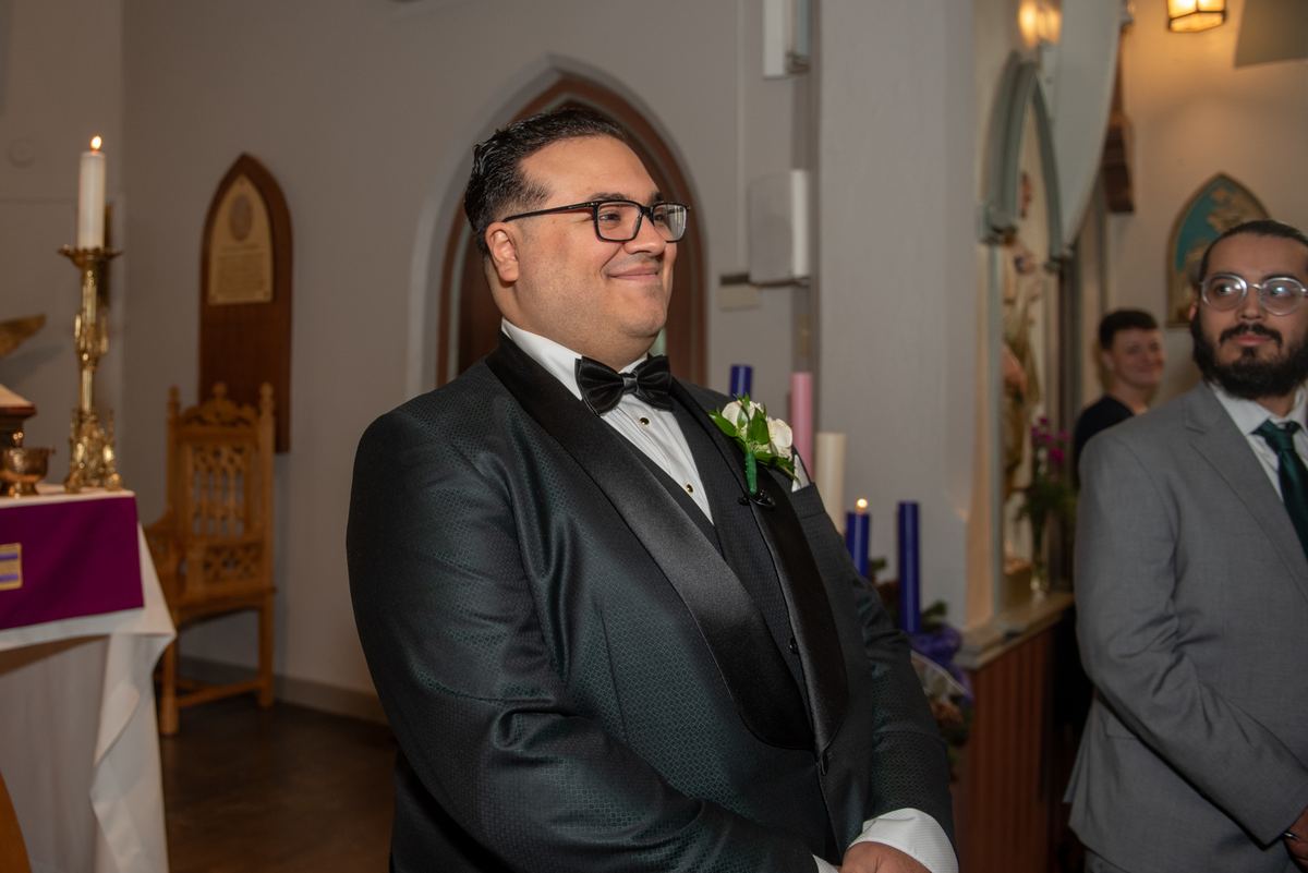 A groom in a dark tuxedo with a boutonnière smiles at his bride during a candle-lit church ceremony, surrounded by guests.