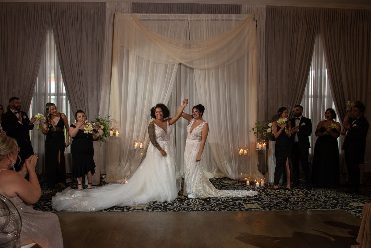 Two brides raise their hands in triumph as guests cheer, surrounded by romantic candlelit ambiance and natural light.