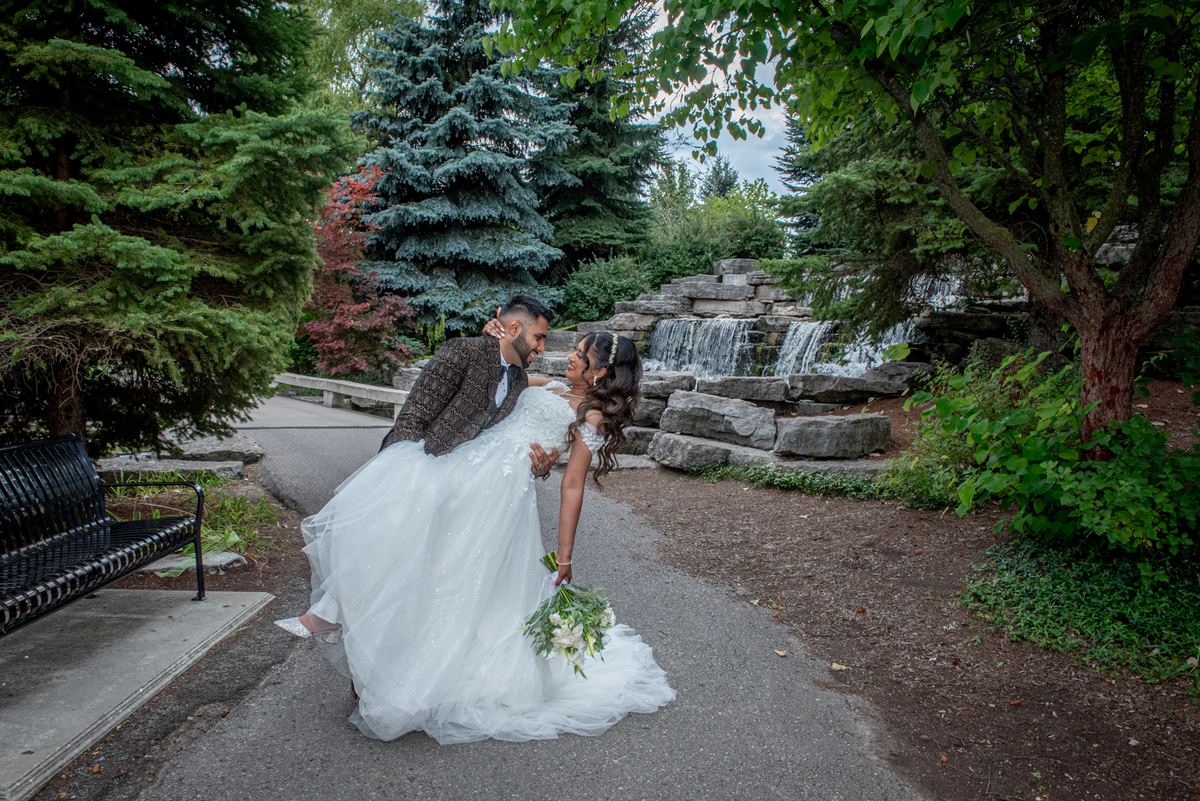 A bride and groom share an intimate moment in a garden, surrounded by greenery and a stone waterfall.