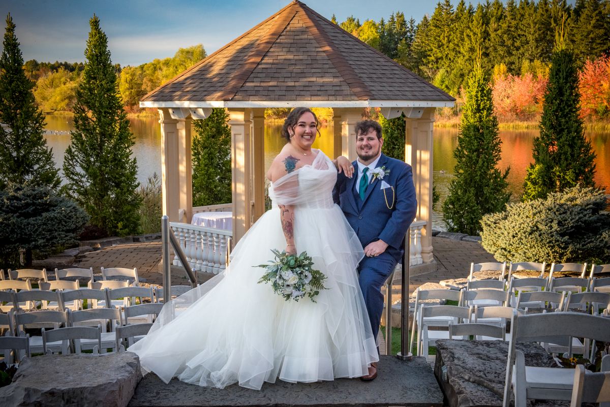 A bride and groom stand joyfully beneath a gazebo by a lake, surrounded by autumn leaves and empty chairs.