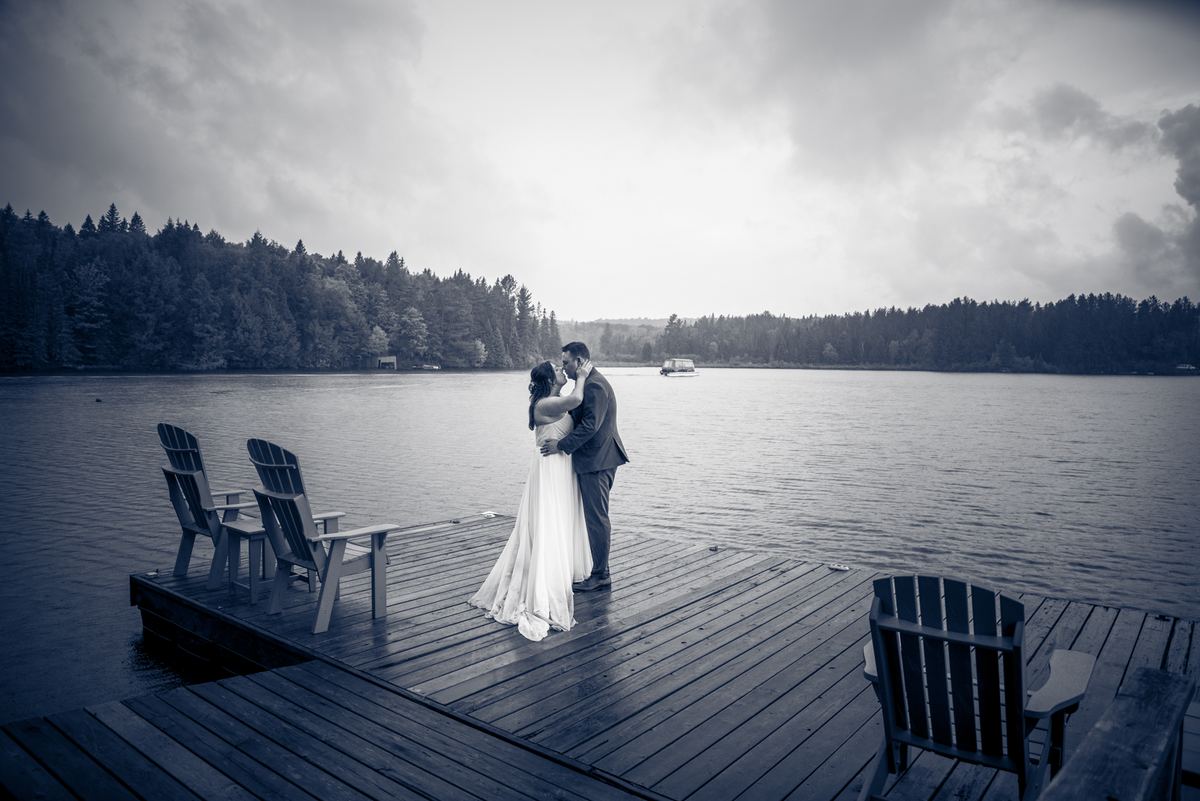 A bride and groom share a kiss on a wooden dock beside a calm lake, framed by empty Adirondack chairs under an overcast sky.