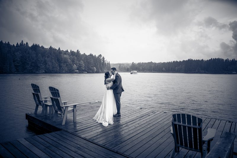 A bride and groom share a kiss on a wooden dock beside a calm lake, framed by empty Adirondack chairs under an overcast sky.
