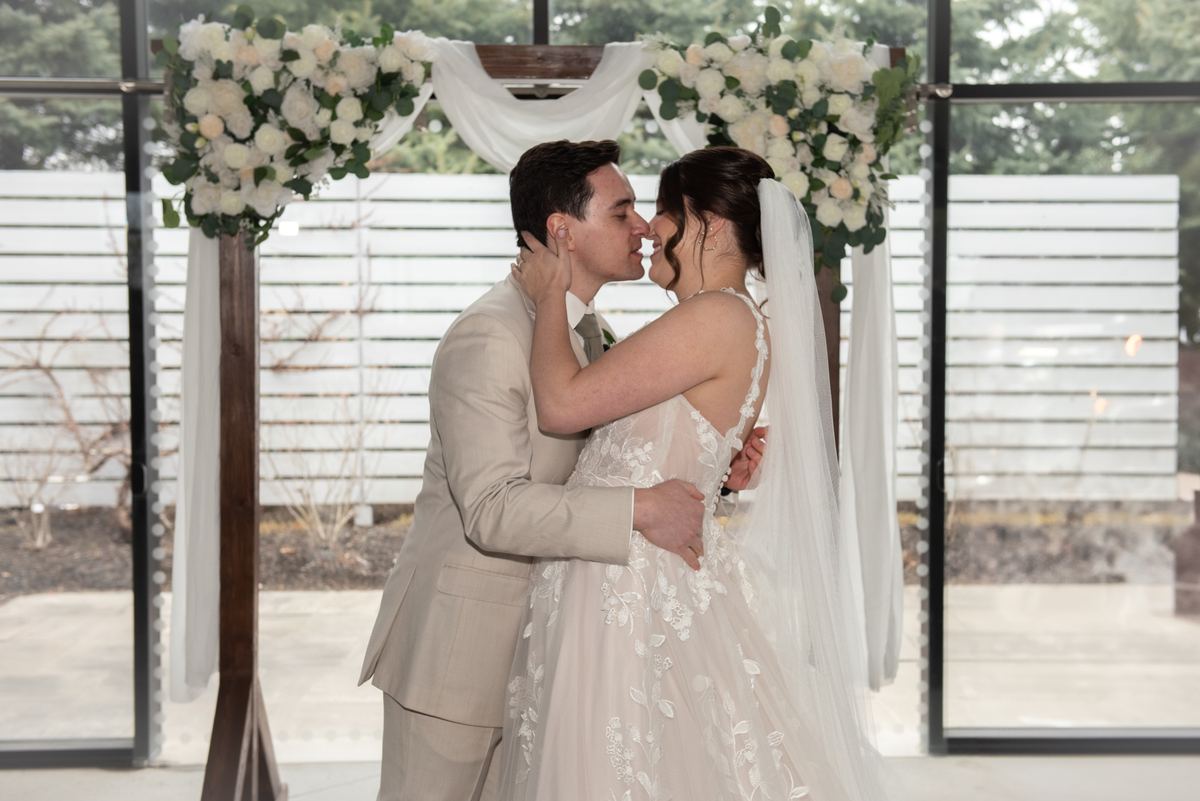 A bride and groom kiss beneath a floral arch decorated with white blooms, illuminated by soft natural light.