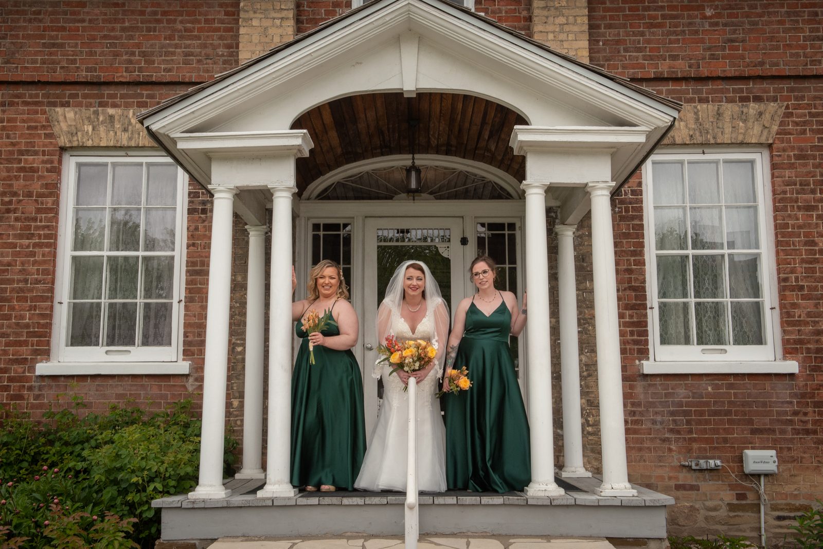 Three women in white and green dresses smile with autumnal bouquets on a porch under a covered portico.