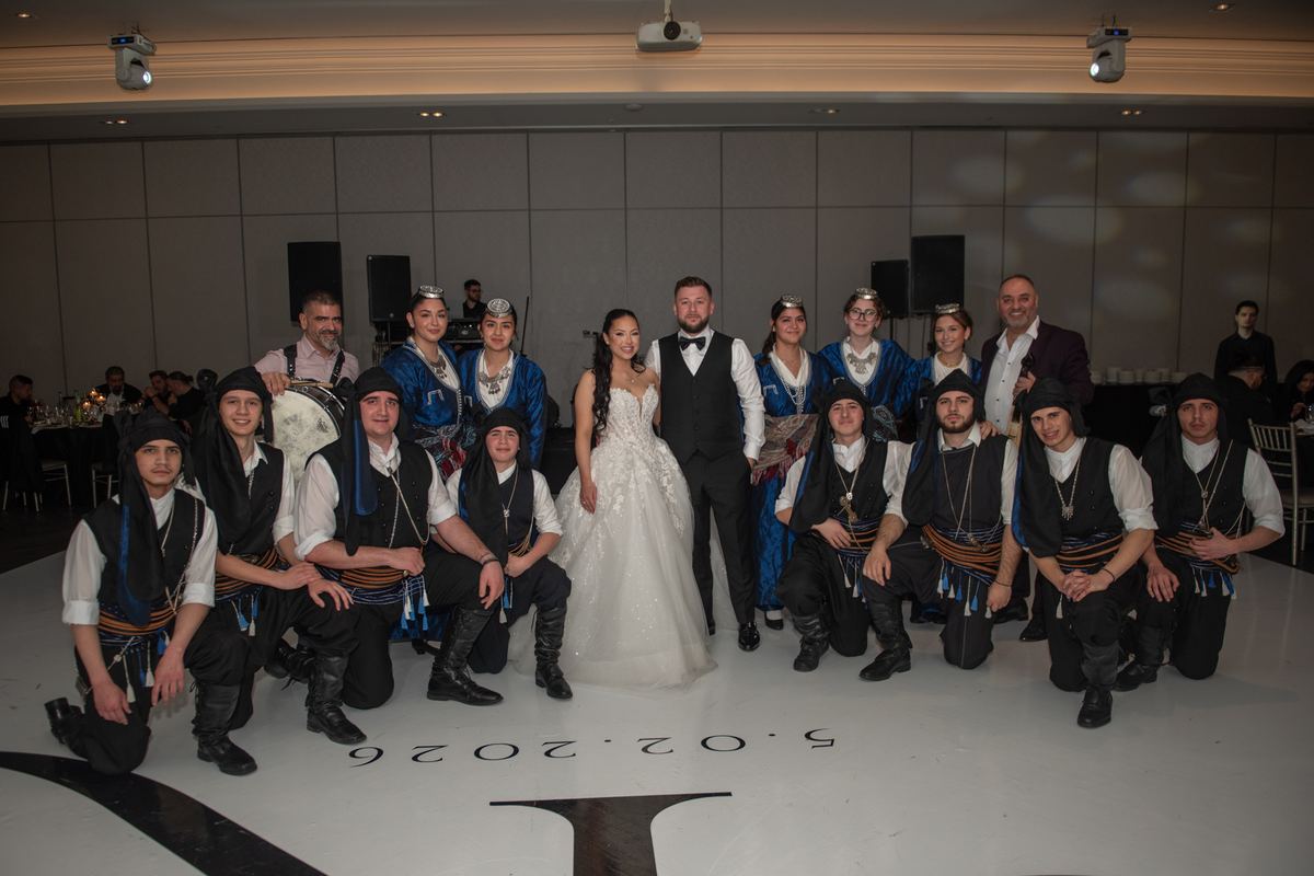 A bride and groom stand centre stage surrounded by a folk ensemble in blue costumes and black vests under bright stage light.
