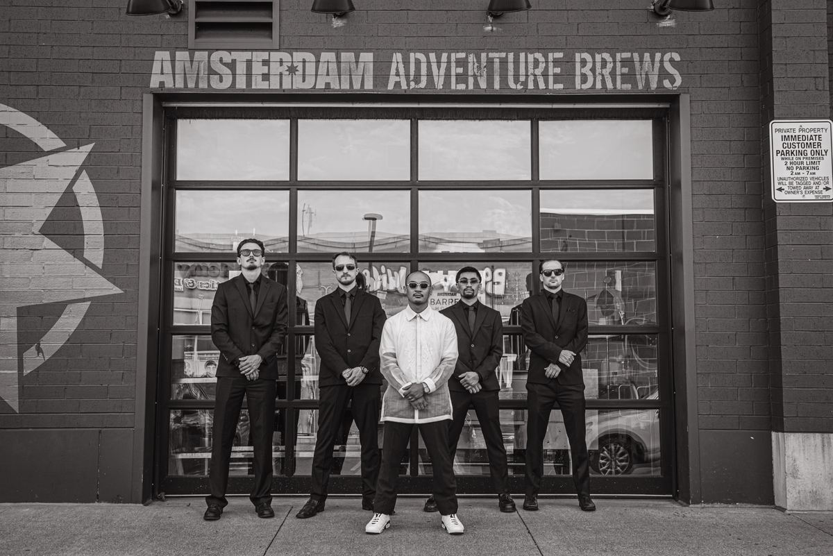 Five men in dark suits and sunglasses stand confidently before a brick building, reflected in its large windows.