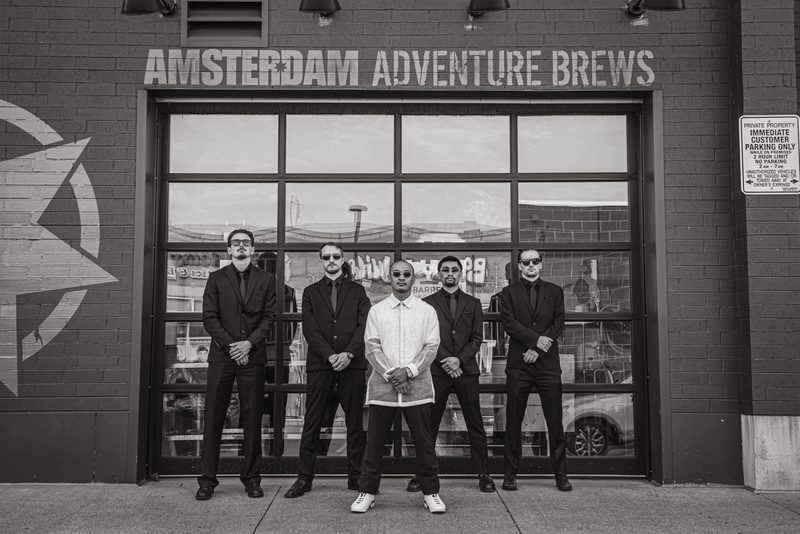 Five men in dark suits and sunglasses stand confidently before a brick building, reflected in its large windows.