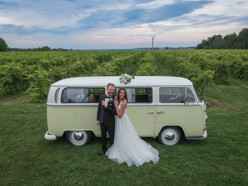 A newlywed couple smiles beside a vintage VW camper van in a vineyard at dusk, glowing softly under the fading light.