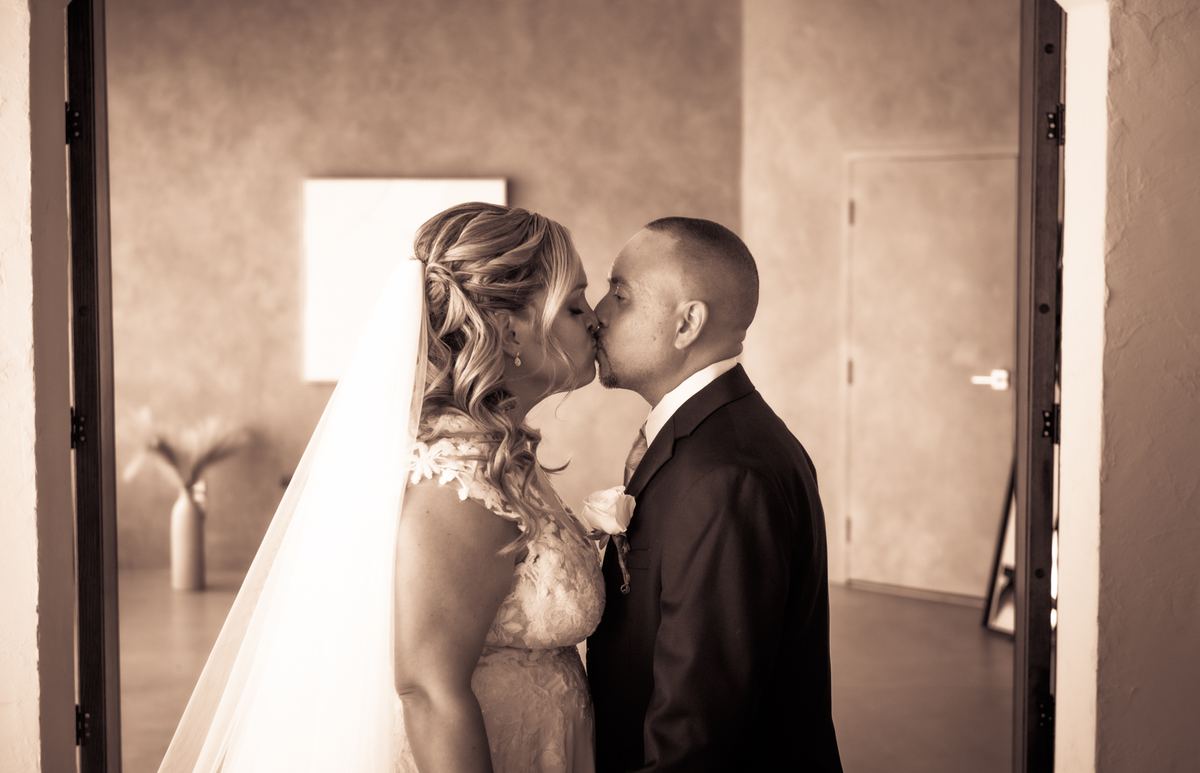A bride and groom kiss tenderly in an open doorway, their profiles illuminated by soft directional light.