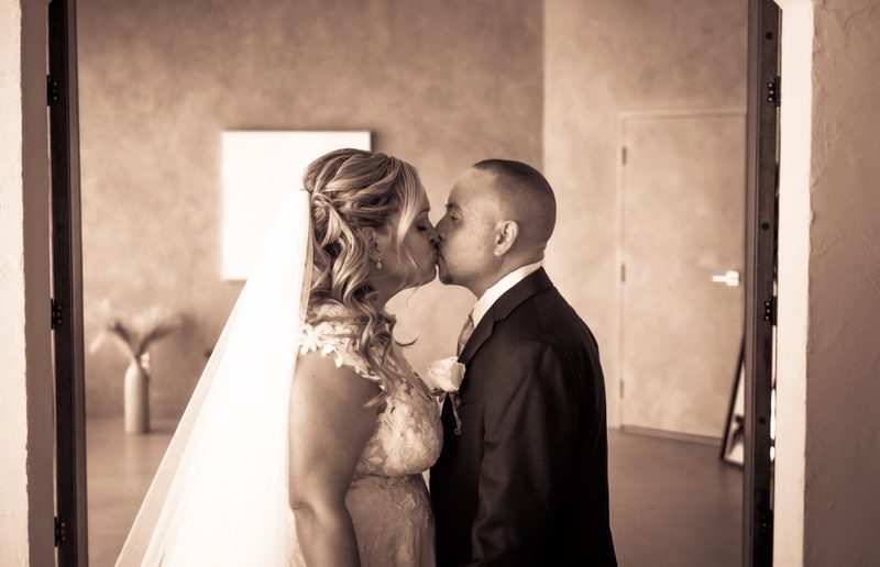 A bride and groom kiss tenderly in an open doorway, their profiles illuminated by soft directional light.
