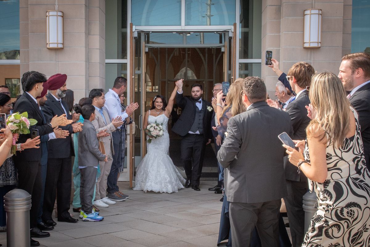 A joyful bride and groom exit a modern church with arms raised as guests applaud, phones out, in bright daylight.