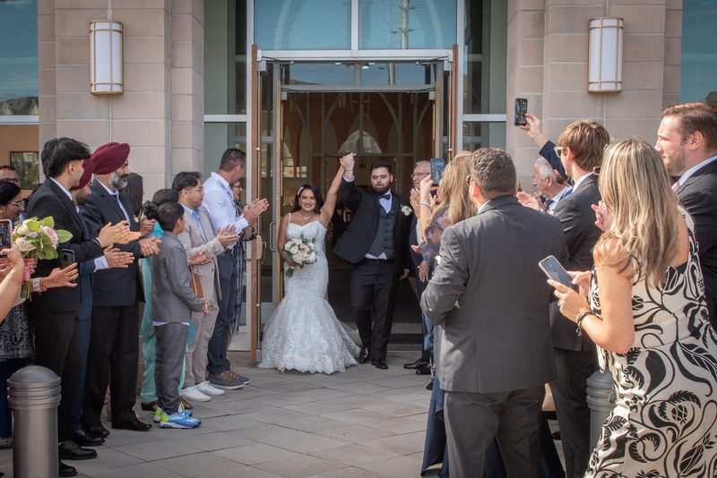 A joyful bride and groom exit a modern church with arms raised as guests applaud, phones out, in bright daylight.