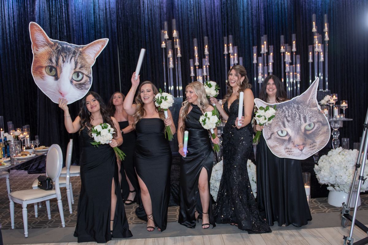 A bridal party in black gowns holds cat face cutouts and white flowers, illuminated by candlelight at a reception.