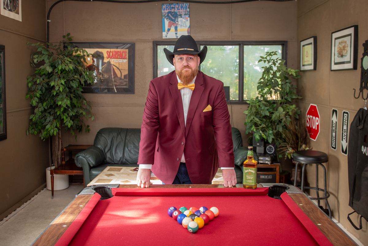 A man in a maroon suit and cowboy hat stands by a red pool table, hands resting on its edge, in a room with eclectic decor.