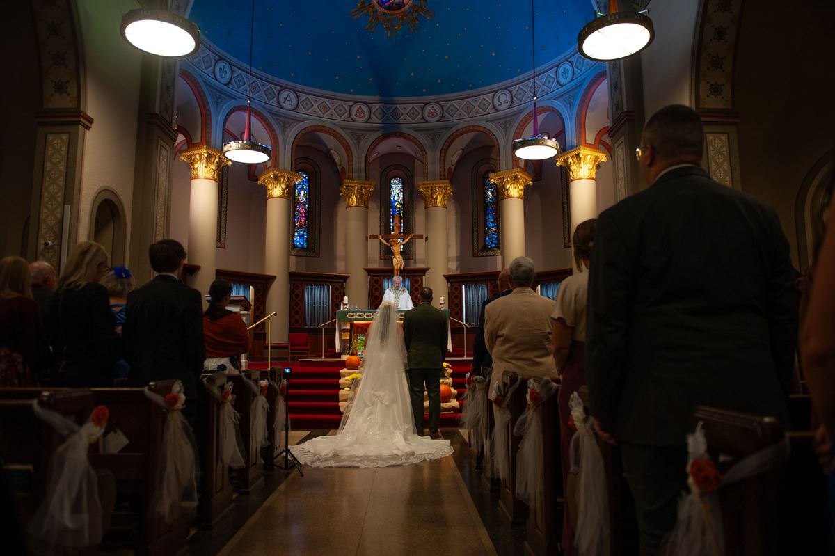 A bride in a white gown and groom in a dark suit walk down the aisle toward an altar under a crucifix, with guests watching.