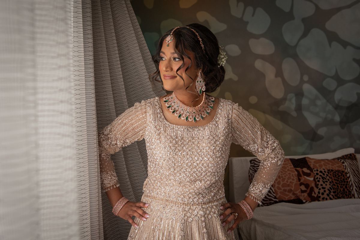 A bride in an ornate cream lehenga with beaded details and traditional jewelry stands by a window bathed in soft light.